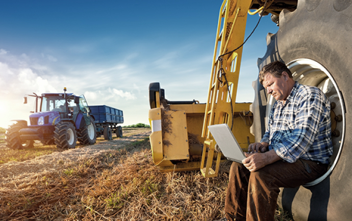 Farmer sitting on tractor tire using laptop to bank.